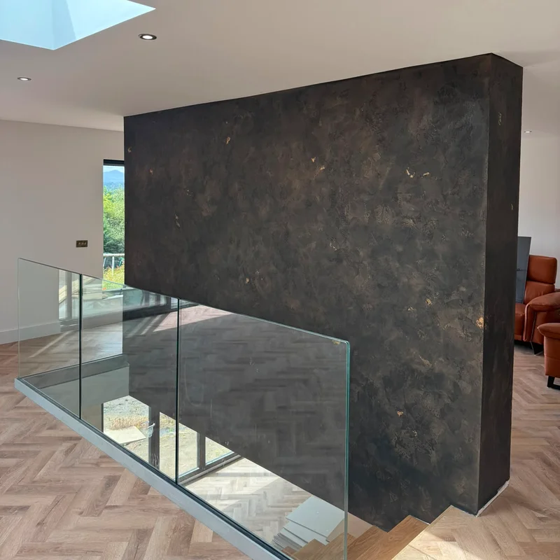 A freestanding landing wall in dark, textured metallic Venetian plaster flecked with gold leaf, behind a glass balustrade