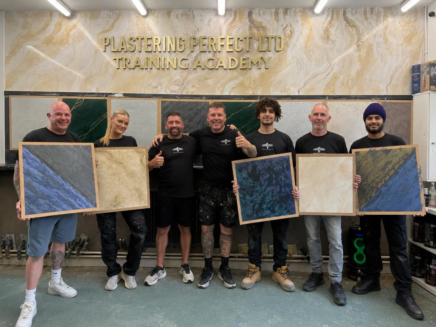 Seven trainees in front of the Plastering Perfect LTD Training Academy sign, each holding a Venetian plaster sample board they made on the course.