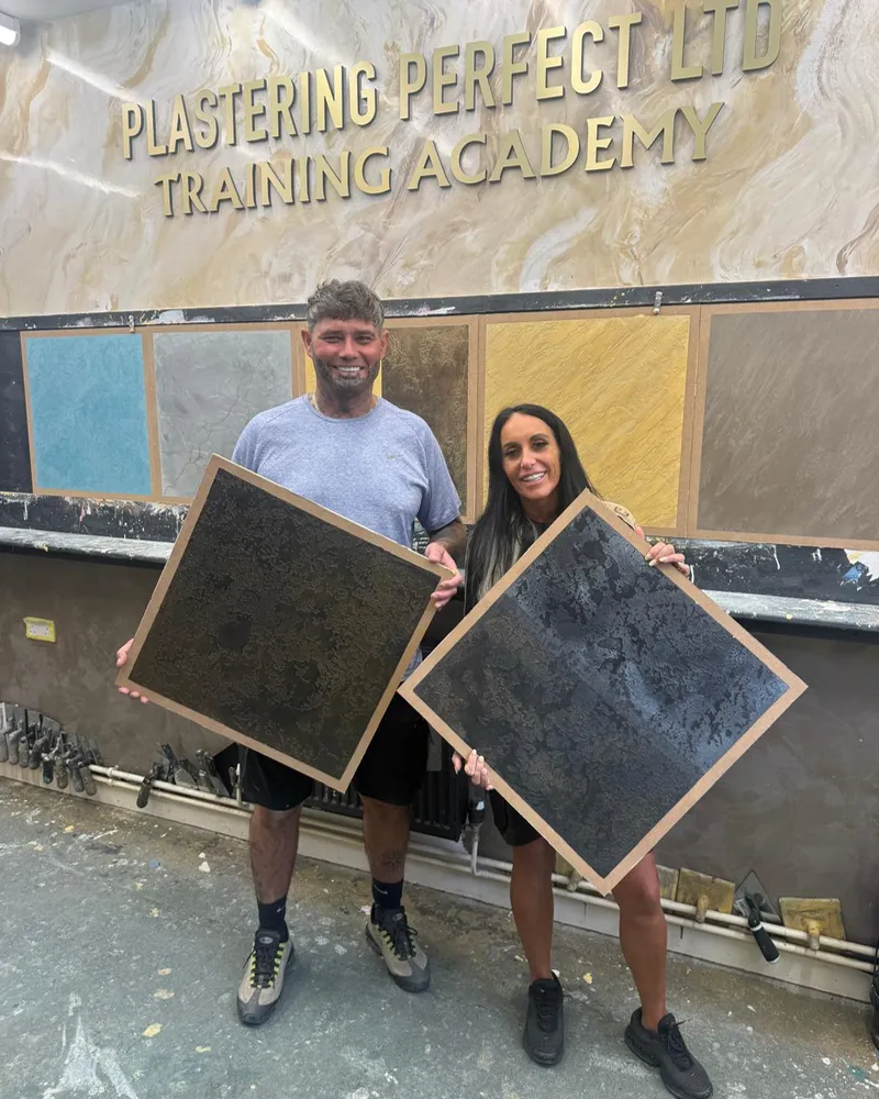 Two trainees under the Training Academy sign, each holding a dark Venetian plaster sample board they made.