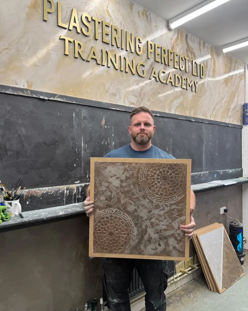A trainee holding a sample board with a raised, textured decorative plaster finish, in front of the Training Academy sign.